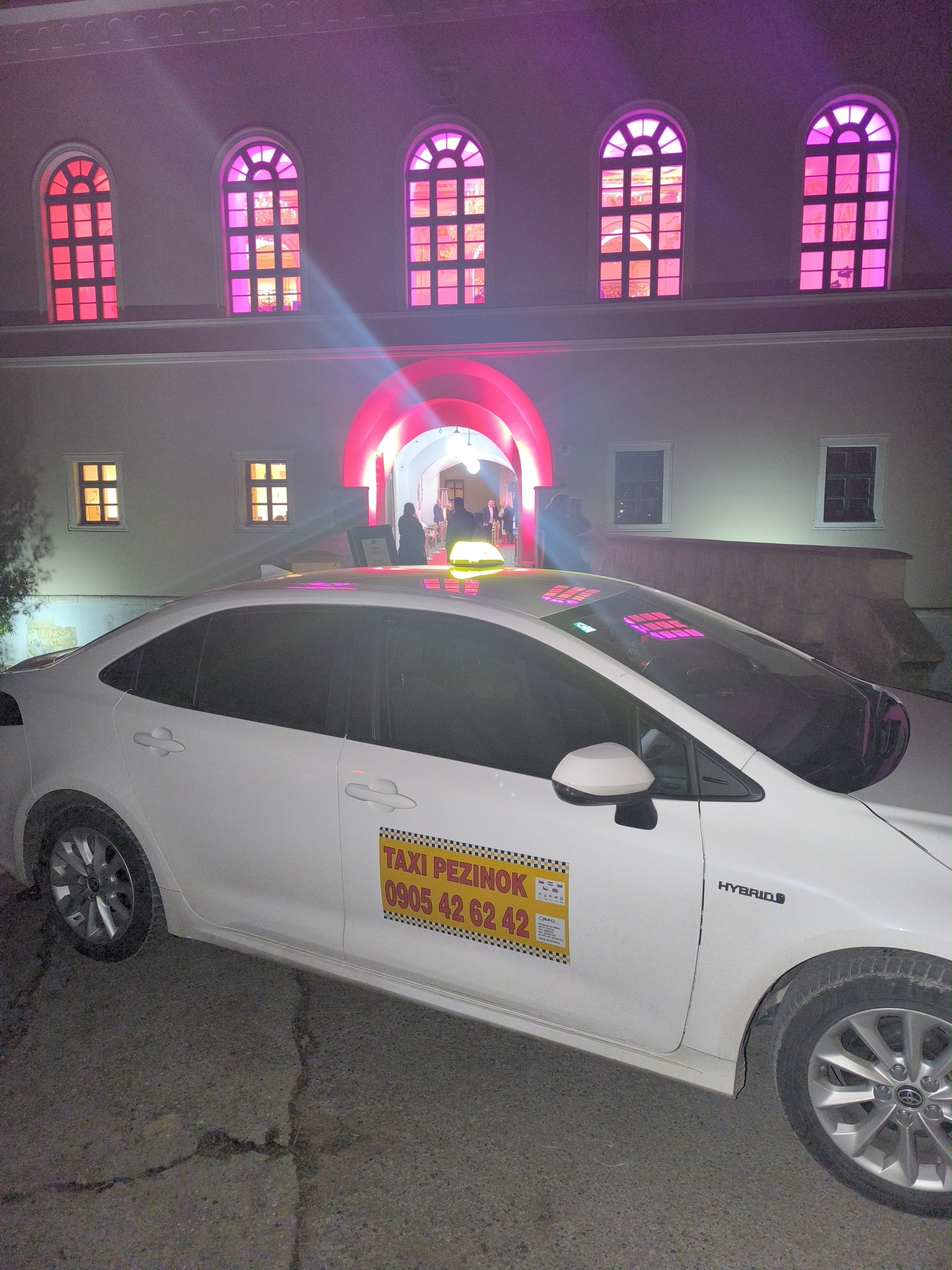 Biele taxi Toyota Corolla s taxametrom a strešným svetlom pred osvetlenou budovou v noci