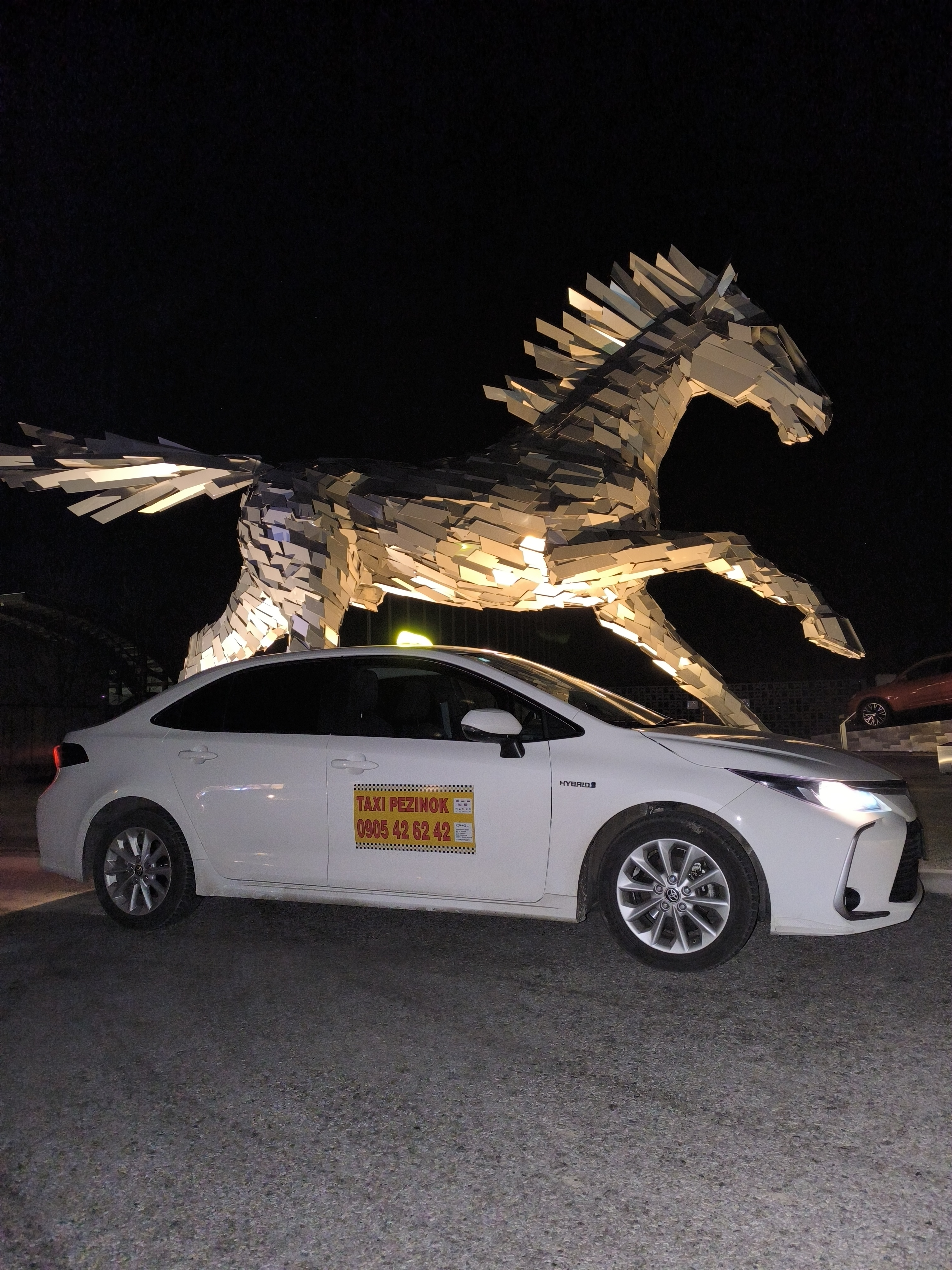 Biele hybridné taxi Toyota Corolla Taxi Pezinok pri nočnej soche koňa
